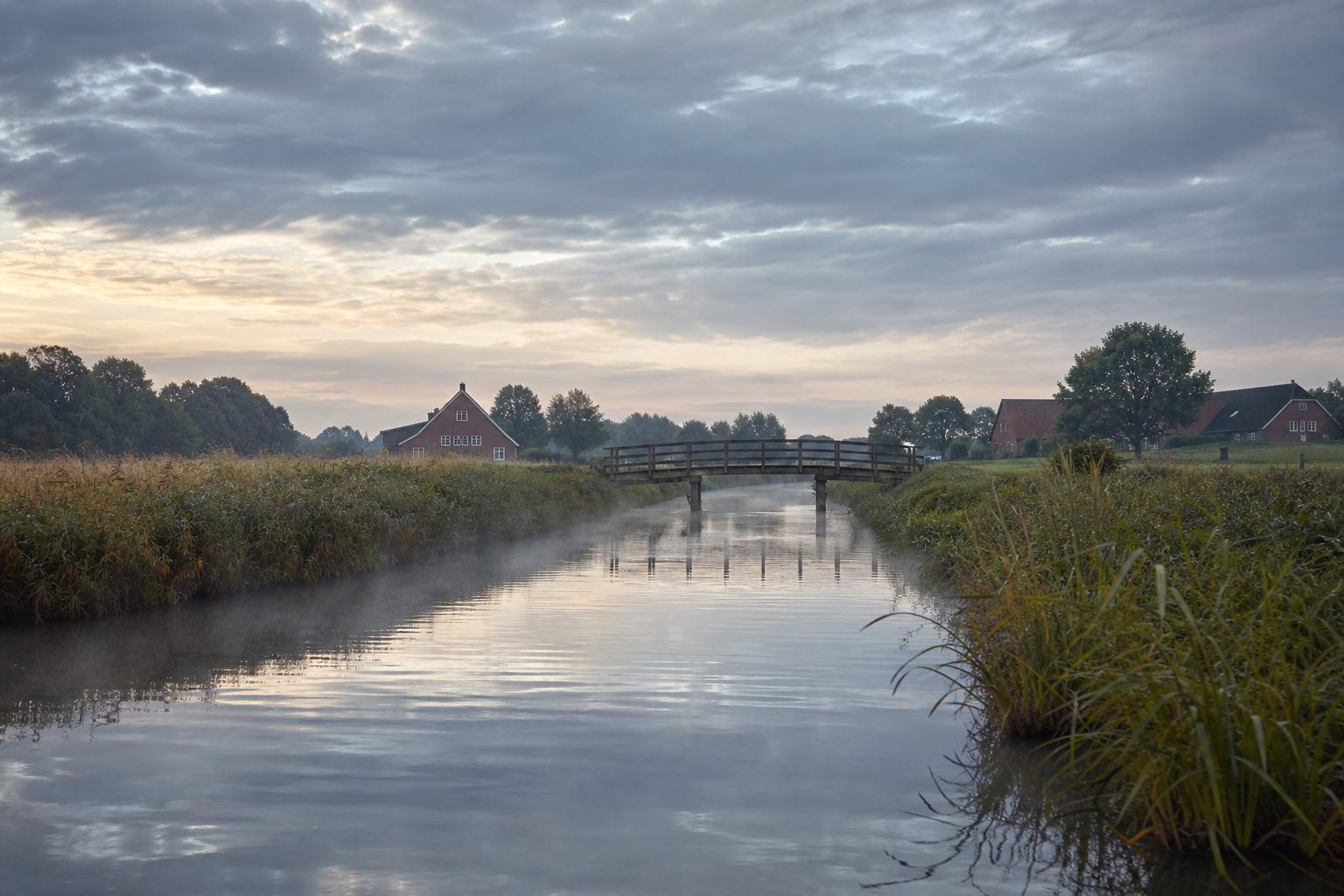 Schmaler Kanal mit Schilf und Brücke unter niedrigem Himmel